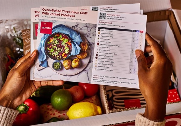 Hands holding a Gousto recipe card above a box of fresh vegetables and lemons